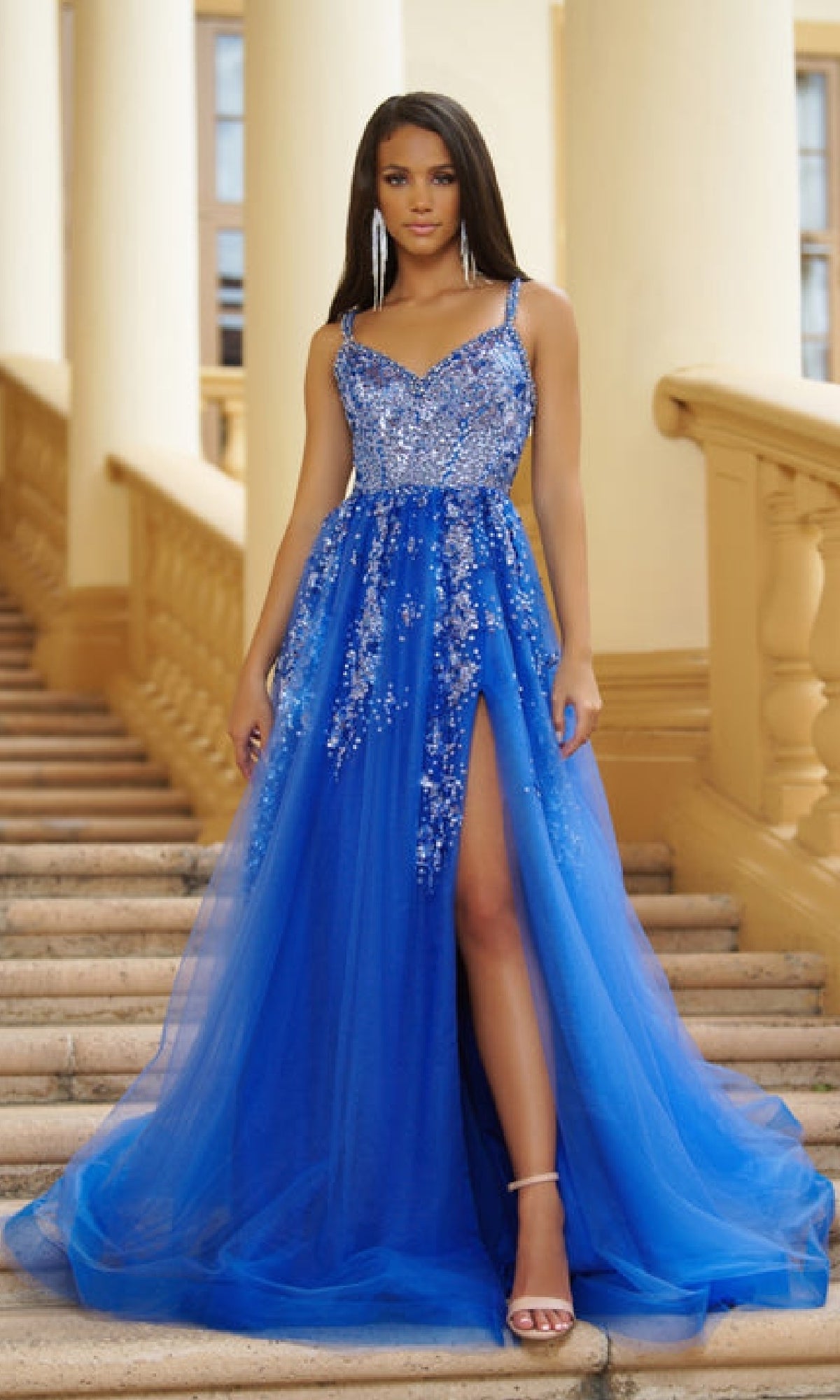 A woman in a Sparkly Long Prom Ball Gown: Ava Presley 28280, featuring bright blue sequins, sparkling details, and a high slit, stands on outdoor steps with columns in the background.